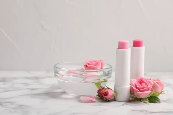 Lip balms, bowl with water and roses on white marble table, space for text