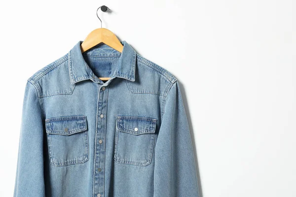 Hanger with denim shirt on white wall, space for text