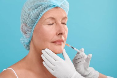 Doktor açık mavi arka planda yaşlı kadına dudak iğnesi yapıyor. Estetik ameliyat