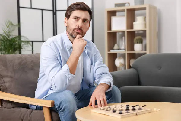 Thoughtful man playing checkers in armchair at home