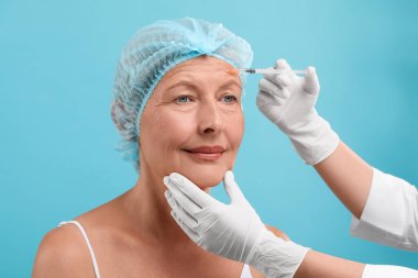 Doktor açık mavi arka planda yaşlı kadına yüz enjeksiyonu yapıyor. Estetik ameliyat
