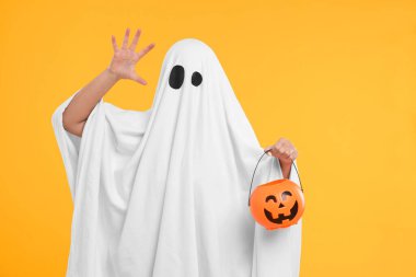 Beyaz hayalet kostümlü kadın sarı arka planda balkabağı kovası tutuyor. Cadılar Bayramı