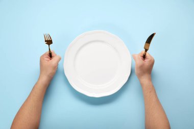 Çatal bıçaklı ve boş tabaklı bir adam açık mavi masada, üst tarafta.