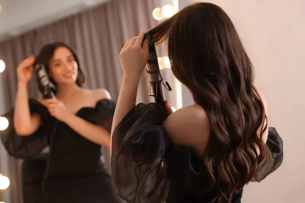 Woman using curling hair iron near mirror at home, selective focus