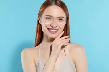 Beautiful young woman with sun protection cream on her face against light blue background