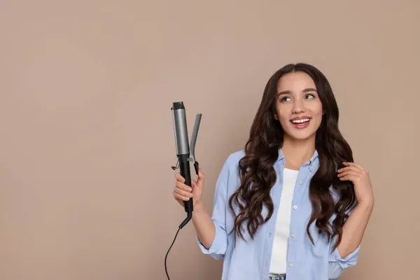 Happy woman with curling hair iron on beige background. Space for text
