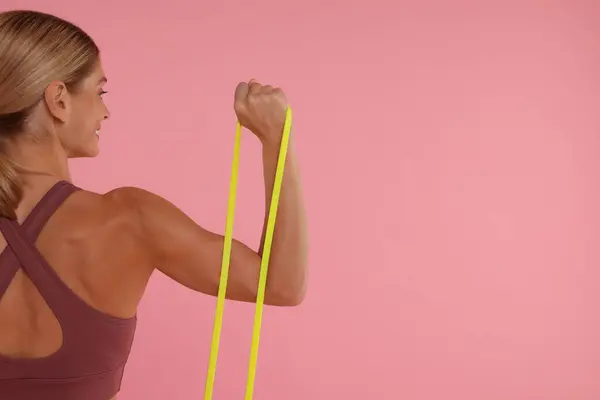 Woman exercising with elastic resistance band on pink background, back view. Space for text