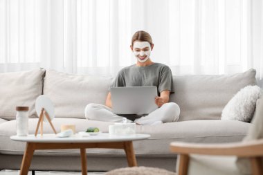 Maskeli genç bir kadın evdeki kanepede dizüstü bilgisayar kullanıyor. Spa tedavileri