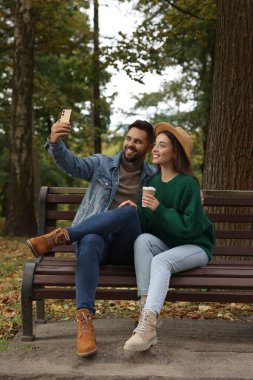 Mutlu genç çift sonbahar parkında ahşap bankta selfie çekiyor.