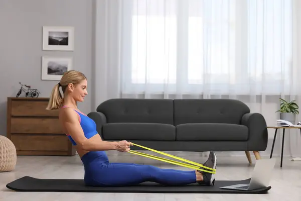 Fit woman doing exercise with fitness elastic band near laptop on mat at home