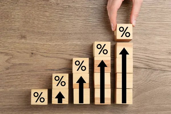 Mortgage rate rising illustrated by upward arrows. Woman putting cube with percent sign near other ones on wooden table, top view