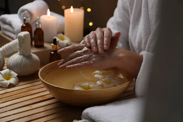 Woman applying scrub onto her hands in spa, closeup. Bowl of water and flowers on wooden table