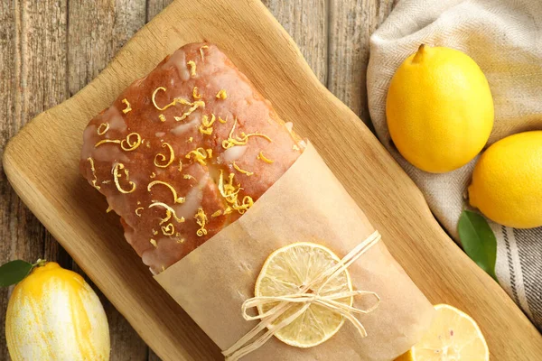Wrapped tasty lemon cake with glaze and citrus fruits on wooden table, flat lay