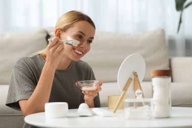 Evde aynanın önüne yüz maskesi takan genç bir kadın. Spa tedavileri