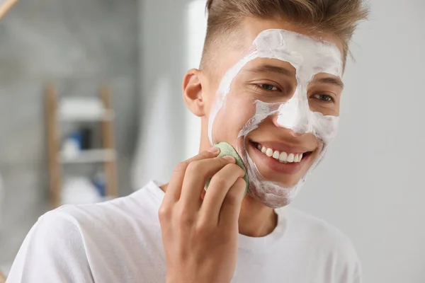 Happy young man washing off face mask with sponge in bathroom