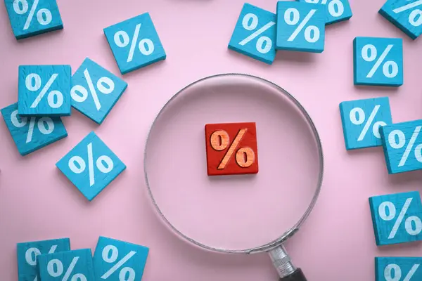 Best mortgage interest rate. Red wooden square with percent sign among light blue ones on pink background, flat lay. View through magnifying glass