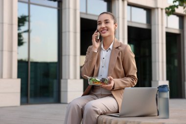 Salata konteynırlı mutlu iş kadını öğle yemeği sırasında dışarıda bankta akıllı telefonla konuşuyor.