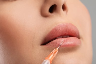 Genç bir kadın açık gri arka planda dudak enjeksiyonu yaptırıyor, yakın plan. Estetik ameliyat