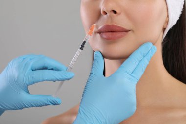 Doktor açık gri arka planda genç bir kadına yüz enjeksiyonu yapıyor, yakın plan. Estetik ameliyat