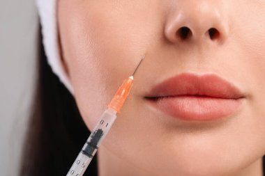 Genç bir kadın açık gri arka planda yüz enjeksiyonu yaptırıyor, yakın plan. Estetik ameliyat