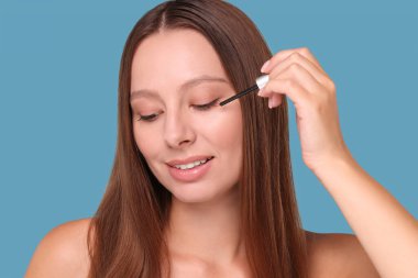 Açık mavi arka planda kirpiklere serum süren güzel bir kadın.