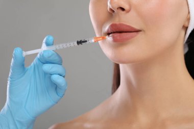 Doktor açık gri arka planda genç bir kadına dudak iğnesi yapıyor, yakın plan. Estetik ameliyat