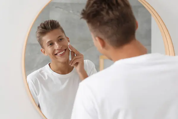 Handsome young man cleaning face with cotton pad near mirror in bathroom