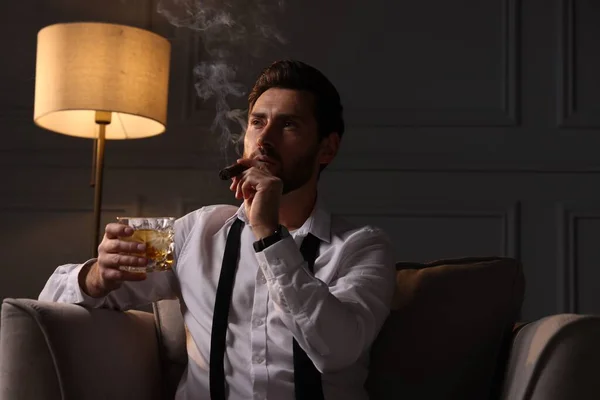 Handsome man with glass of whiskey smoking cigar at home. Space for text