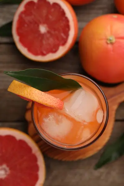 Tasty grapefruit drink with ice and leaf in glass surrounded by fresh fruits on wooden table, flat lay