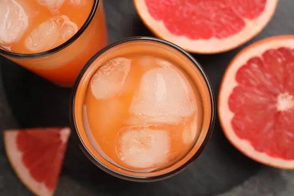 Tasty grapefruit drink with ice in glasses and fresh fruits on dark table, flat lay