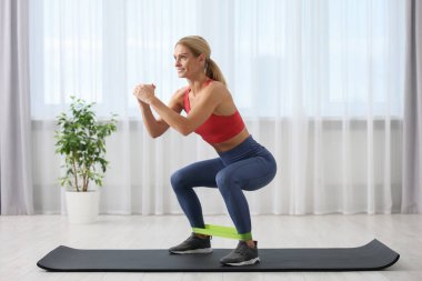 Athletic woman doing squats with fitness elastic band on mat at home clipart