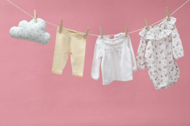 Farklı bebek kıyafetleri ve bulut şekilli yastık pembe arka planda çamaşır ipinde kuruyor.