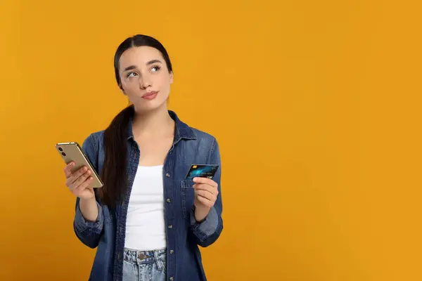 Confused woman with credit card and smartphone on orange background, space for text. Debt problem