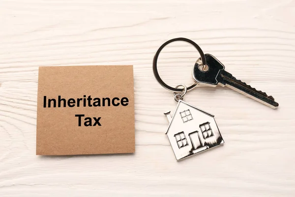 Inheritance Tax. Card and key with metallic key chain in shape of house on white wooden table, flat lay
