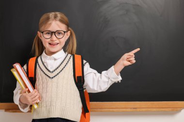 Sırt çantası ve kitaplarıyla gözlüklü mutlu öğrenci tahtaya yakın bir yeri işaret ediyor.