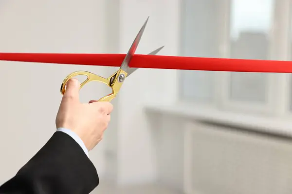 Woman cutting red ribbon with scissors indoors, closeup