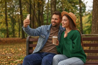 Mutlu genç çift sonbahar parkında ahşap bankta selfie çekiyor.