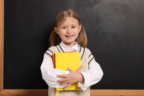 Happy schoolgirl with backpack and books near blackboard