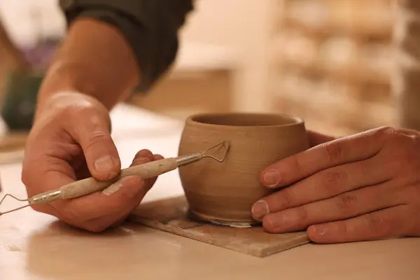 Clay crafting. Man making bowl with sculpting tool at table in workshop, closeup