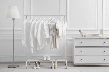 Beyaz duvarın yanında farklı şık kadın kıyafetleri, ayakkabıları, lambası ve şifoniyeri var.