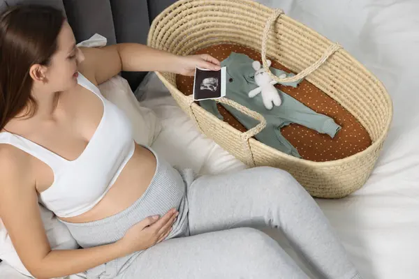 Pregnant woman with ultrasound picture of baby and basket on bed, above view
