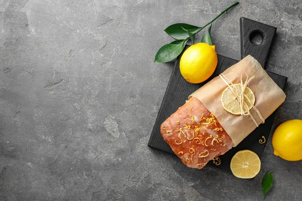 Wrapped tasty lemon cake with glaze and citrus fruits on grey textured table, flat lay. Space for text