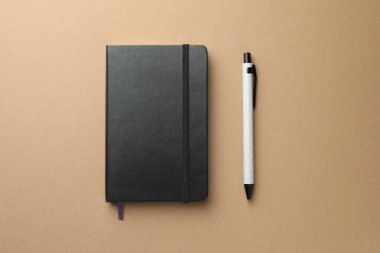 Açık kahverengi zemin üzerinde kapalı siyah defter ve kalem, üst görünüm