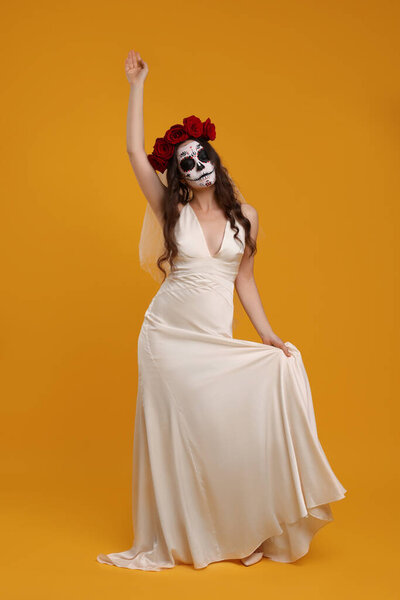 Young woman in scary bride costume with sugar skull makeup and flower crown posing on orange background. Halloween celebration