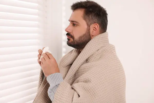 Sick man wrapped in blanket with tissue indoors. Cold symptoms