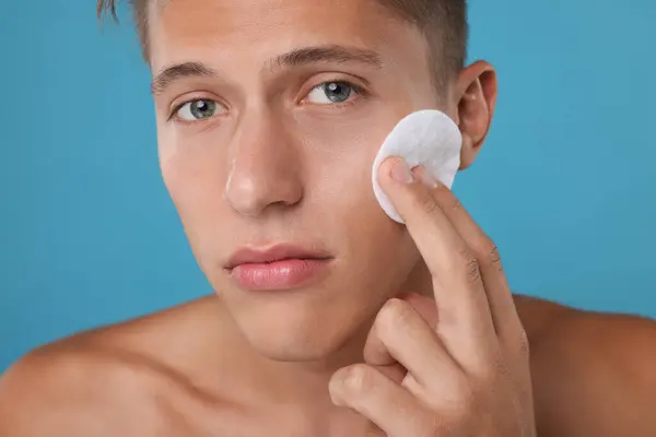 Handsome man cleaning face with cotton pad on light blue background