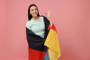 Alman bayrağı taşıyan mutlu genç bir kadın pembe arka planda iyi niyet gösterisi yapıyor.