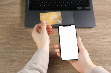 Çevrimiçi ödeme. Kredi kartı ve akıllı telefon kullanan bir kadın, ahşap masanın yanında boş ekranlı, yakın plan.
