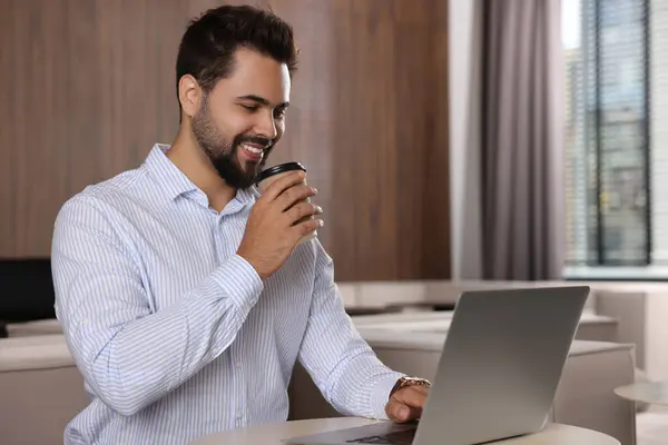 Happy young man with paper cup of coffee working on laptop at table in office