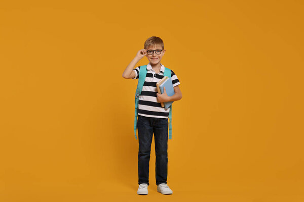 Happy schoolboy in glasses with backpack and books on orange background